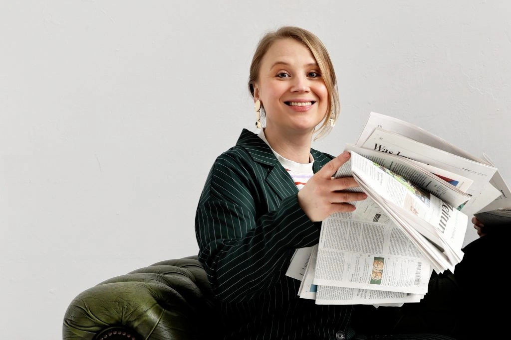 Marie Franz, Gründerin der PR-Agentur Pressegold Berlin, lächelt mit einem Stapel Zeitungen auf einem grünen Ledersessel