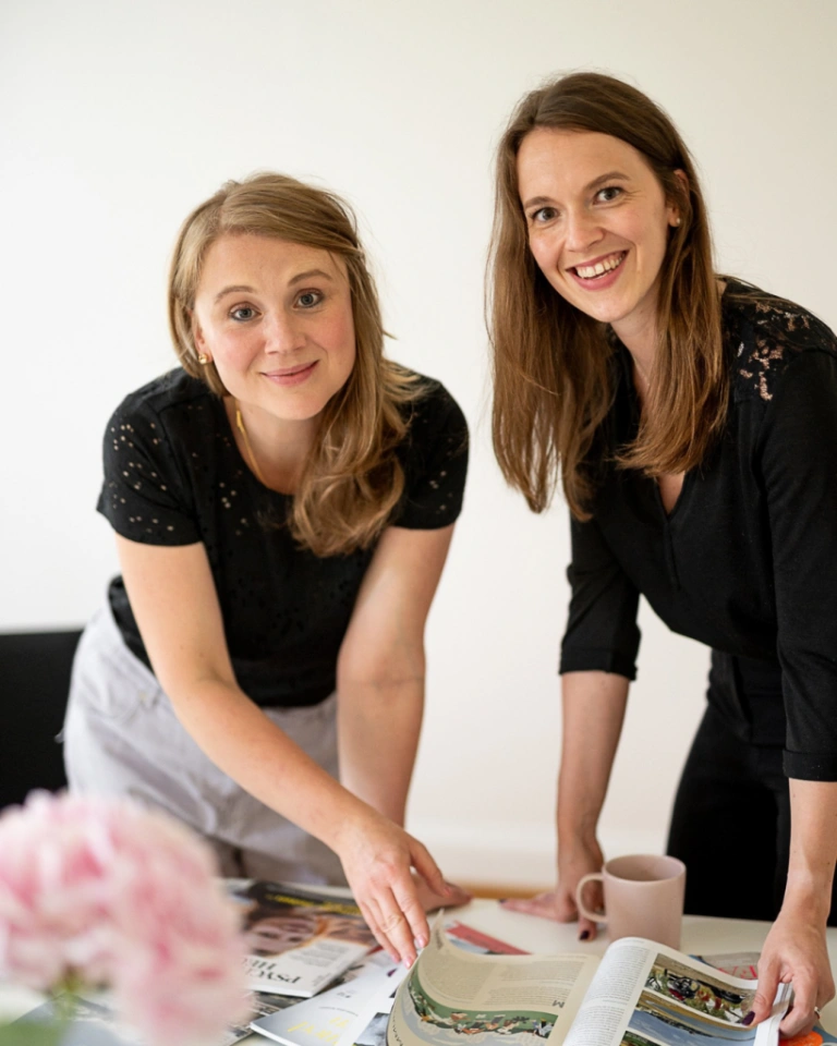 Marie Franz und Sophia Lock, Gründerinnen der PR-Agentur Pressegold Berlin, beugen sich lächelnd über einen Tisch mit Magazinen